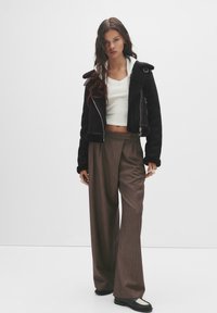Giacca nera in pelle ecologica con zip argentata, maglietta bianca a collo a V corta e pantaloni marroni a gamba larga, caratterizzati da una sottile texture e pieghe.