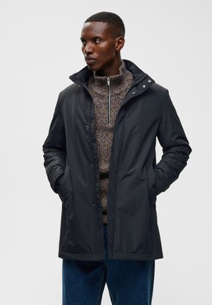Homme portant une veste noire zippée sur un pull marron à motifs, les mains dans les poches, regardant à gauche sur un fond uni.