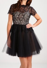 Svart klänning med en spetstopp med blommönster, korta ärmar och en lager av tylskjol. Strukturerad tyg med en figursydd midja.