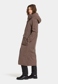 Bruine geïsoleerde lange jas met capuchon, ritssluiting, zijzakken en een rechte snit. Gecombineerd met zwarte jeans en zwarte schoenen.