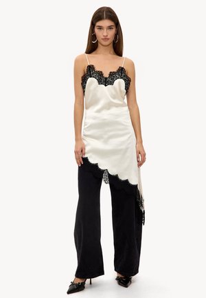 Femme portant une robe nuisette en satin blanc avec bordure en dentelle noire, par-dessus un pantalon noir à jambes larges et des escarpins noirs à bouts pointus, se tenant devant un fond blanc.