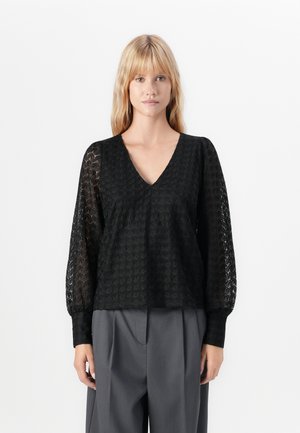 Woman wearing black lace long-sleeve blouse with V-neck and loose gray high-waisted trousers, standing against white background.