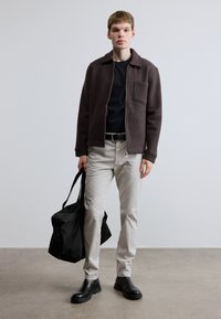 Krátka tmavohnedá bunda na zips cez čierne tričko, svetlosivé nohavice, čierne topánky a držiaca čiernu tašku. Hladká tkanina s golierom a manžetami na rukávoch.