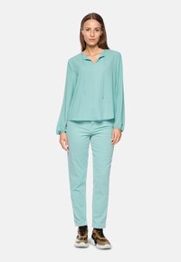 Mintgroene blouse met lange mouwen en capuchon, gecombineerd met bijpassende mintgroene slim-fit broek en bruin-witte sneakers met luipaardprintaccenten.