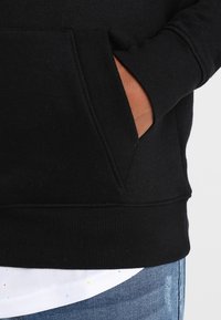 Felpa nera con polsini a coste e una tasca frontale, realizzata in materiale morbido. Sotto indossa una camicia bianca con punti colorati.