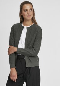 Cardigan vert foncé, texturé avec des points en relief, avec un col rond et une ouverture à l'avant. Porté par-dessus un haut blanc avec un pantalon noir.