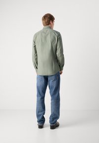 Cămașă button-up de culoare verde închis cu guler răspândit, combinată cu jeansi drepti de culoare albastru deschis. Cămașa are mâneci lungi și o textură netedă.