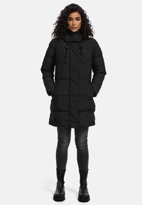 Abrigo acolchado negro con cuello alto, cremallera frontal y cordones. Tiene bolsillos laterales y llega hasta la rodilla, combinado con jeans oscuros y botas negras.
