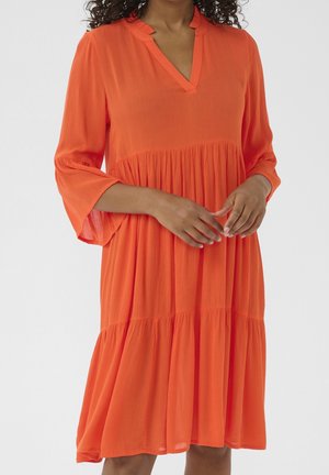 Femme portant une robe ample orange vif, longueur genoux, avec manches trois-quarts et col en V, sur un fond uni.