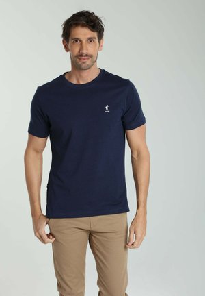Homme portant un t-shirt bleu marine à col rond avec un petit logo girafe et un pantalon beige, debout devant un fond gris uni.