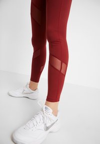 Gambe che indossano leggings rossi con pannelli in rete e scarpe Nike bianche con loghi swoosh argentati su uno sfondo bianco.