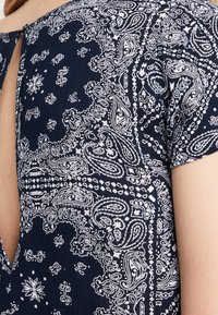 Gros plan sur un tissu bleu marine avec un motif cachemire blanc et une ouverture en forme de trou de serrure près du haut du dos, montrant une partie de l'épaule et du bras d'une personne.