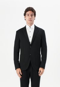 Schwarzer Blazer mit schlanker Passform, Reverskragen, einreihigem Knopfverschluss und zwei Vorderaschen, kombiniert mit einem weißen Hemd mit schwarzen Knöpfen.