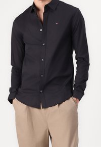 Maglietta nera a maniche lunghe con colletto a camicia, caratterizzata da un piccolo logo bianco, rosso e blu sul petto e una texture liscia.