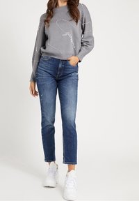 Grauer Pullover mit Perlenverzierungen, kombiniert mit hellblauen, abgenutzten Jeans und weißen Turnschuhen. Lässiger, entspannter Schnitt und modernes Design.