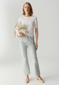 Top bianco a maniche corte con orlo a scallop, jeans dritti verde chiaro, ballerine beige, che tiene una borsa tote panna.
