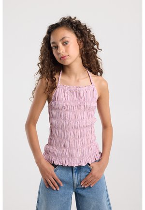 Jonge vrouw met krullend haar, gekleed in een lichtroze gerimpelde haltertop en een blauwe spijkerbroek, staand met handen in de zakken tegen een effen achtergrond.