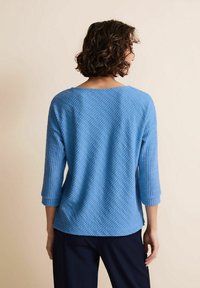 Blå, strukturerad tröja med en lös passform, trekvartslång ärm och vid halsringning, med ett diamantmönster. Bärs med mörka byxor.