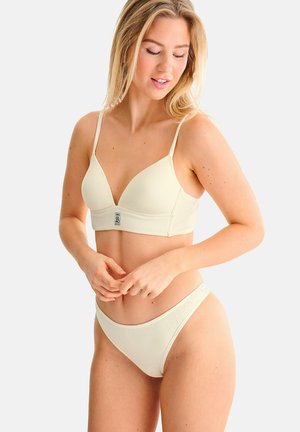 Femme portant un soutien-gorge côtelé crème et des culottes taille haute assorties, regardant vers le bas avec les mains près de la taille, sur un fond clair uni.