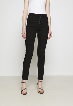 Femme portant un jean skinny noir taille haute avec fermeture à boutons à l'avant et des sandales noires à talons hauts et brides, debout sur un fond uni.