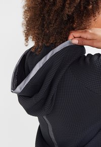 Haut à capuche noir texturé au design ajusté, avec une doublure perforée grise à l'intérieur de la capuche et des accents réfléchissants discrets.