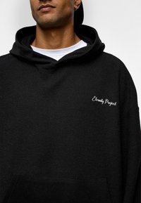 Sweatshirt à capuche noire en tissu texturé, avec une poche avant et un logo brodé "Eternity Project" sur la poitrine.