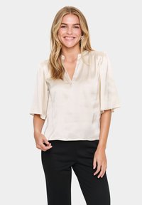 Blusa in satin beige con maniche a sbuffo corte, scollo a V e vestibilità rilassata; indossata con pantaloni neri. Texture morbida e lucentezza elegante.