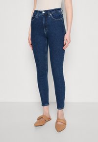 Jeans skinny de cintura alta en azul oscuro, con una textura de denim suave y cinco bolsillos, combinados con zapatos de tiras cruzadas en color tan.