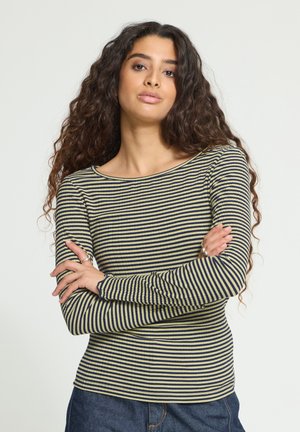 Jeune femme aux longs cheveux bouclés portant un haut à manches longues rayé jaune et noir et un jean bleu, bras croisés, expression neutre.