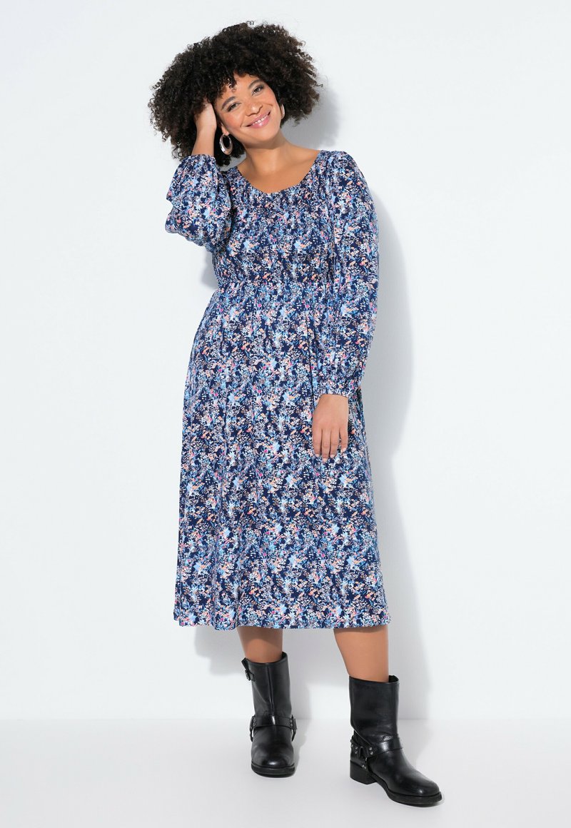 Frau mit lockigem Haar, die ein blaues Midikleid mit Blumenmuster und schwarze Knöchelstiefel trägt, lächelnd mit der Hand auf dem Kopf vor weißem Hintergrund.