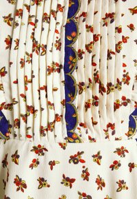 Tissu blanc avec petit imprimé floral rouge, jaune et marron, avec plis verticaux et bordure décorative bleu marine festonnée aux bords dorés.