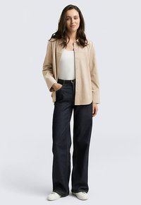 Chemise beige à manches longues, jean bleu foncé à jambes larges, et baskets blanches. La chemise est dotée de boutons et d'une coupe décontractée. Texture lisse.