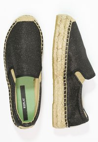 Chaussures noires tissées à enfiler avec une finition texturée et des accents élastiques sur les côtés. Semelle de style jute avec des coutures visibles et une étiquette de semelle intérieure verte.