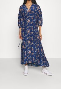 Robe maxi fleurie bleue avec décolleté en V, manches trois-quarts, ceinture ajustable à la taille et silhouette fluide ; associée à des baskets blanches.