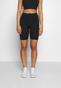 Černé vysokým pasem zdůrazněné cyklistické šortky s přiléhavým střihem a hladkým povrchem, v kombinaci s bílými sportovními botami. Minimalistické logo na straně.