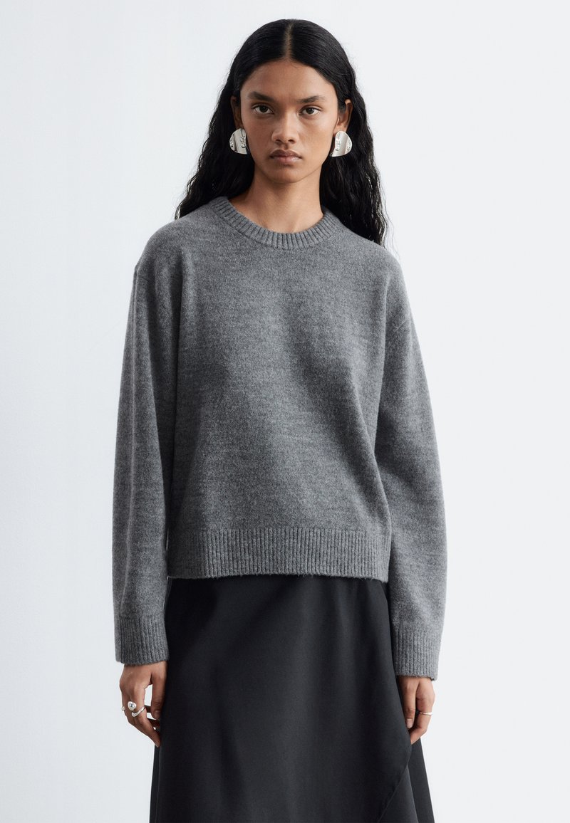 Femme aux longs cheveux noirs portant de grandes boucles d’oreilles argentées, un pull en tricot gris et une jupe noire, debout devant un fond clair uni.
