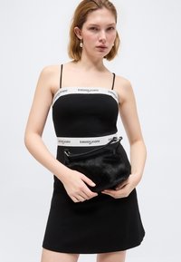 Γυναίκα που φοράει μαύρο αμάνικο φόρεμα με λευκές λωρίδες "tommy jeans", κρατώντας μια μικρή μαύρη γούνινη τσάντα σε απλό λευκό φόντο.