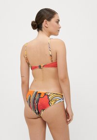 Bikini set s viacfarebným abstraktným vzorom, s červeným vrchom s nastaviteľnými ramienkami a oranžovými spodkami, obidva s detalmi šitia.