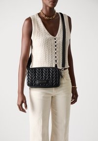 Borsa a tracolla in pelle nera trapuntata con motivo testurizzato, hardware argento e tracolla regolabile, abbinata a un top in maglia leggera e pantaloni crema.