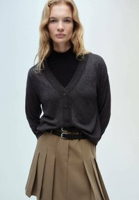 Cardigan gri închis din lână, cu decolteu în V, cinci nasturi și margini striate, asociat cu o fustă plisată bej și o curea neagră. Textura netedă a lânei.
