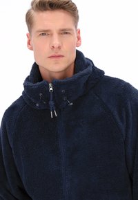 Granatowa polarowa kurtka z wysokim kołnierzem, przednim zamkiem błyskawicznym i ściągaczem. Miękka tekstura z prążkowanymi detalami i solidnymi srebrnymi elementami.