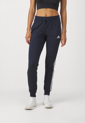Vrouw draagt marineblauwe Adidas trainingsbroek met witte strepen aan de zijkanten, gecombineerd met witte sneakers en een zwart crop topje.