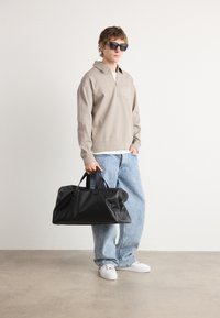 Hanorac bej cu fermoar și logo CK, blugi lejere albaștri, adidași albi și o geantă de voiaj din piele neagră. Modelul poartă ochelari de soare închiși la culoare.
