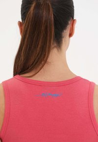 Vaaleanpunainen hihaton paita, jossa on pyöreä kaula-aukko. Takana sininen brodeerattu teksti, näkyviä ompeleita ja sileä kangastuntuma.