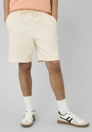 Homme portant un short beige clair à cordon, des chaussettes blanches hautes et des baskets blanches avec des touches noires, debout devant un fond uni.
