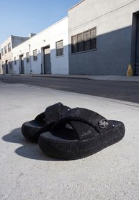 Sandales à plateforme noires pailletées avec larges brides croisées reposant sur un trottoir urbain ensoleillé près de bâtiments industriels.