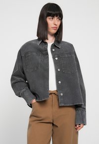 Gray denim jacket with a collared neck, two chest pockets, and silver buttons. Worn over a white top and paired with brown pants.