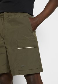 Personne portant un short Nike vert olive avec une taille élastique, main dans la poche latérale et poche cargo cousue avec bordure réfléchissante.