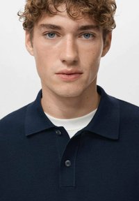 Granatowa koszulka polo o fakturowanej tkaninie, z dwoma guzikami przy kołnierzu, noszona na białej koszulce z okrągłym dekoltem, o prostym kroju.