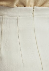 Tissu texturé blanc des pantalons, avec une fermeture éclair dissimulée et de subtils plis au niveau de la taille. Fini lisse.